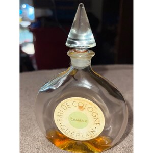 Puede incluir: Un frasco de perfume de vidrio transparente con cuerpo redondo y un tapón de vidrio cónico y puntiagudo. El frasco presenta una etiqueta circular con el texto EAU DE COLOGNE GUERLAIN CHAMADE. La base contiene un líquido de color ámbar.