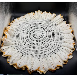 Könnte beinhalten: Runder, cremefarbener Häkeldoily mit einem spiralförmigen Zentrum und einem gewellten Rand. Der Rand ist mit einer dünnen gelben Bordüre versehen. Der Doily ist handgefertigt und hat ein offenes, durchbrochenes Design.