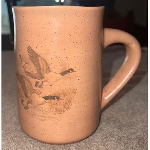 May include: A cylindrical ceramic mug with a speckled light orange finish. The exterior features a dark brown illustration of two geese in flight over a marshy area. The mug has a thick, curved handle on the right side and a smooth, rounded rim.
