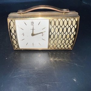 May include: Vintage gold-toned travel alarm clock with a white face and a decorative lattice design on the right side. The clock has a handle and the word "SWIZA" is printed on the face. The clock is rectangular and has a classic design.