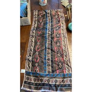 May include: A rectangular scarf with a patterned design. The scarf features a combination of colors, including black, pink, blue, and cream. The design includes floral and geometric patterns. The scarf has a fringed edge.