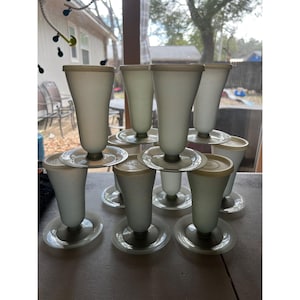 May include: A collection of vintage white frosted glass parfait glasses with matching lids and saucers. The glasses have a flared shape and are stacked on top of each other. The lids and saucers are a light beige color.