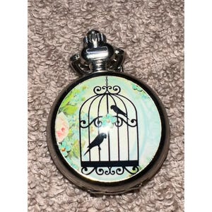 May include: A round silver-toned pocket watch case featuring a decorative insert with a black silhouette of two birds inside a birdcage. The background of the insert displays a light blue and green floral pattern with soft pink accents.