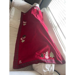 Puede incluir: Una manta de color rojo intenso con un borde a cuadros y motivos navideños bordados que incluyen muñecos de nieve, árboles de hoja perenne y copos de nieve blancos. La tela tiene una textura suave y está colocada sobre una superficie blanca.