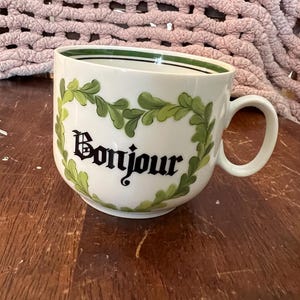 May include: White ceramic mug with a green leaf design and the word "Bonjour" in black script. The mug has a rounded shape with a handle and a green stripe around the rim. The mug is on a wooden surface.