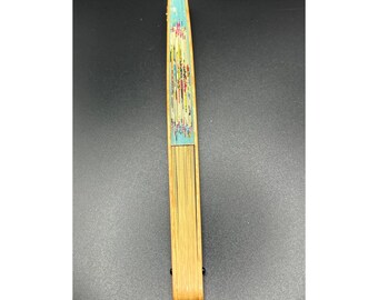 May include: A closed, rectangular hand fan with a floral design. The fan's handle is made of light-colored wood, and the top section features a blue background with colorful flowers. The fan is approximately 10 inches long.
