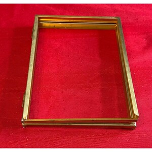 May include: A rectangular, gold-colored metal frame, possibly for a photograph or artwork, is displayed against a red background. The frame has a simple, elegant design with clean lines and a slight sheen.