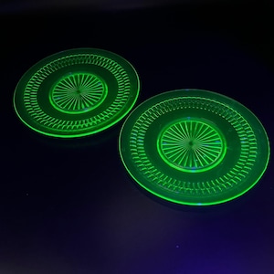 May include: Two round, neon green glass plates with a central starburst design and a decorative outer ring. The plates are illuminated against a dark background, showcasing their vibrant color and intricate details. The plates are likely vintage or antique.