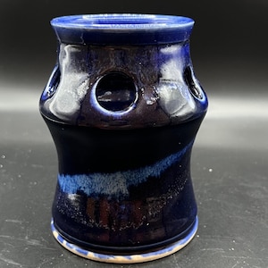 May include: A handcrafted ceramic vase in shades of deep blue with a glossy finish. The vase features a cylindrical shape with a flared base and a series of circular openings around the upper portion. The interior is a lighter blue, creating a striking contrast.