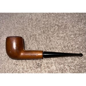 May include: A tobacco pipe with a smooth, medium-brown wooden bowl and a long, tapered black stem. The side of the bowl is stamped with the text LONGCHAMP FRANCE. The pipe features a classic, curved design with a polished finish on the wood.