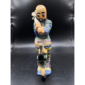 May include: A ceramic figurine depicting a bearded figure with a bald head, wearing a striped outfit in shades of blue, green, yellow, and purple. The figure holds a large, light-colored club over one shoulder while standing upright on a base.