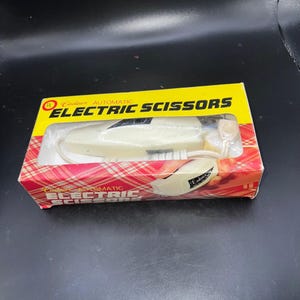 May include: A white Cadaux automatic electric scissors tool housed in a rectangular yellow and red plaid cardboard box. The packaging features the text Cadaux AUTOMATIC ELECTRIC SCISSORS and a UL logo, with a clear plastic window displaying the device.