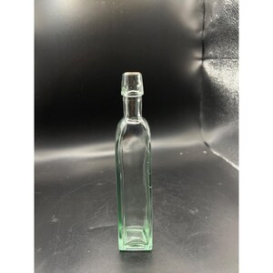 May include: A tall, slender, clear glass bottle with a rectangular base and a narrow neck. The bottle features a distinct, slightly flared rim at the top opening. The glass has a subtle light green tint throughout its structure, and the bottle stands upright.