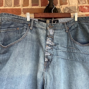 May include: Light blue denim jeans with distinctive stitching and a button fly. The jeans are suspended on a wooden hanger against a brick backdrop. The jeans feature front pockets and a striped detail on the waistband.