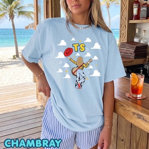 May include: A light blue t-shirt featuring a graphic of a person with long hair playing a guitar, wearing a yellow plaid shirt and cow-print pants, holding a red hat. The text TS is printed above, surrounded by white clouds and musical notes.