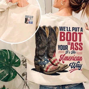 May include: Cream-colored sweatshirt with a graphic of cowboy boots featuring the American flag design. The text on the back reads "We'll Put a Boot in Your Ass, It's The American Way." A smaller graphic on the front says "Brought to you courtesy of the Red, White & Blue."
