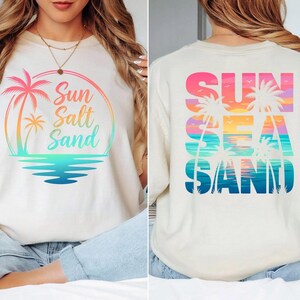 Puede incluir: Camiseta color crema con un gr&aacute;fico colorido. La parte delantera presenta una palmera y las palabras "Sun Salt Sand" en un degradado arco&iacute;ris. La parte trasera muestra las palabras "Sun Sea Sand" con siluetas de palmeras.
