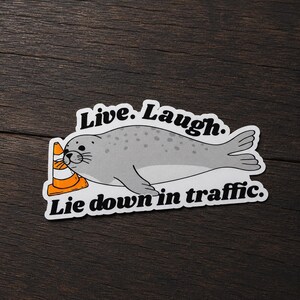 May include: A die-cut sticker featuring a grey spotted seal resting its head against an orange and white traffic cone. The text above the seal reads Live. Laugh. and the text below the seal reads Lie down in traffic. in black font.