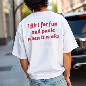 Może przedstawiać: Biała koszulka z czerwonym napisem "I flirt for fun and panic when it works". Koszulka jest w stylu casual, prawdopodobnie wykonana z bawełny i noszona na zewnątrz.