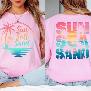 Puede incluir: Camiseta rosa claro con dise&ntilde;o gr&aacute;fico colorido. La parte delantera muestra "Sun Salt Sand" con palmeras y un atardecer. La parte trasera presenta "Sun Sea Sand" en un estilo similar, evocando la playa.