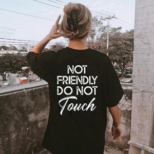 May include: Black oversized t-shirt with the text "NOT FRIENDLY DO NOT Touch" in white. The shirt is worn by a person with their back to the camera. The person is wearing a hair clip and a bracelet.
