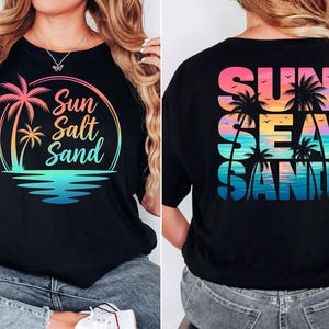 Puede incluir: Camiseta negra con un dise&ntilde;o gr&aacute;fico colorido. La parte delantera presenta una palmera y las palabras "Sun Salt Sand" en un dise&ntilde;o circular. La parte posterior de la camiseta muestra "Sun Sea Sand" con palmeras y p&aacute;jaros.
