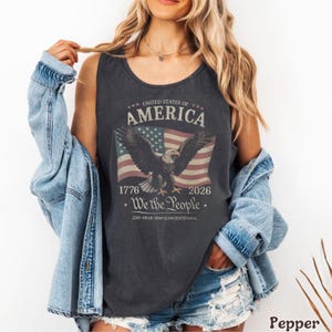 Puede incluir: Una camiseta sin mangas de color gris carbón con un gráfico de un águila calva con las alas extendidas sobre una bandera de los Estados Unidos. El texto dice UNITED STATES OF AMERICA, 1776, 2026, We the People y 250-YEAR SEMIQUINCENTENNIAL.