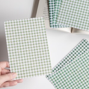 May include: A stack of green and white gingham patterned cards next to an open book. One card is being held by a hand. The cards have rounded corners and a light green and white checkered design. The book pages are visible in the background.