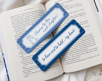 Könnte beinhalten: Zwei blaue und weiße rechteckige Lesezeichen mit Text und Illustrationen auf einem offenen Buch. Eines sagt "Cheers to the next chapter!" und das andere "to have and to hold... my book!"