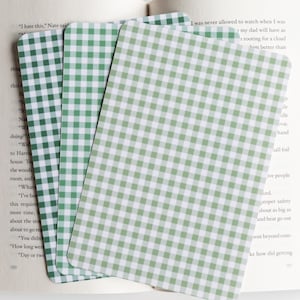 May include: Three rectangular cards with a green and white gingham pattern are fanned out on top of an open book. The cards are a light green color and have rounded corners. The book pages are visible in the background.