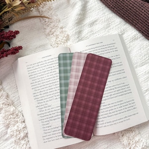 May include: Three rectangular bookmarks with a plaid pattern in shades of sage green, dusty rose, and deep burgundy, resting on the open pages of a book. The bookmarks are thin, flat, and feature a consistent grid design across their surfaces.