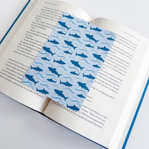 May include: A rectangular bookmark featuring a repeating pattern of dark blue sharks swimming amidst light blue wavy lines. The bookmark is placed on the open pages of a book with printed text, showing page numbers 224 and 225.