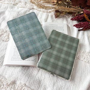 May include: Two rectangular e-reader cases featuring distinct patterns. One case displays a teal background with a subtle grid of small, shimmering holographic stars. The second case features a muted sage green and gray plaid tartan pattern.