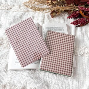 May include: Two rectangular electronic tablet covers featuring a brown and white gingham check pattern. One cover is placed flat on an open book, while the other is positioned slightly overlapping it, showcasing the consistent checkered design.