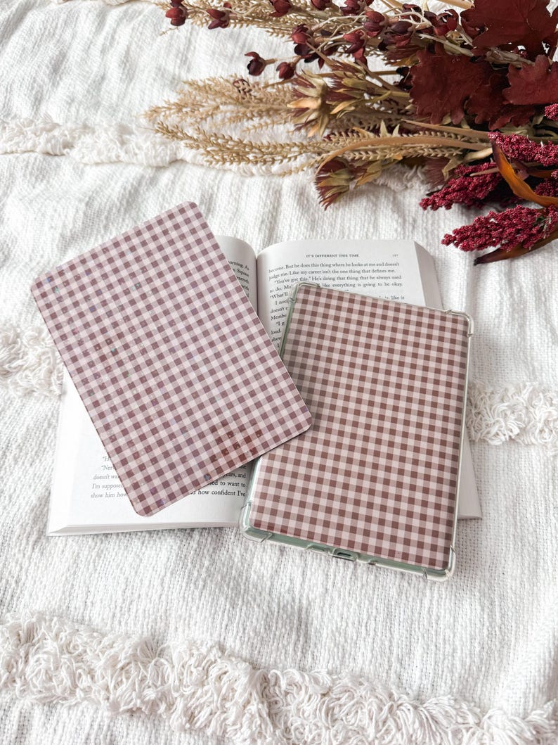 May include: Two rectangular electronic tablet covers featuring a brown and white gingham check pattern. One cover is placed flat on an open book, while the other is positioned slightly overlapping it, showcasing the consistent checkered design.