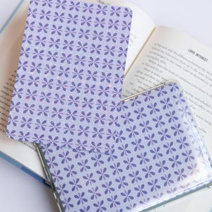 May include: A clear phone case and a matching card with a floral pattern in shades of purple rest on an open book. The case is transparent, showcasing the design beneath. The book's pages are visible, adding context to the scene.