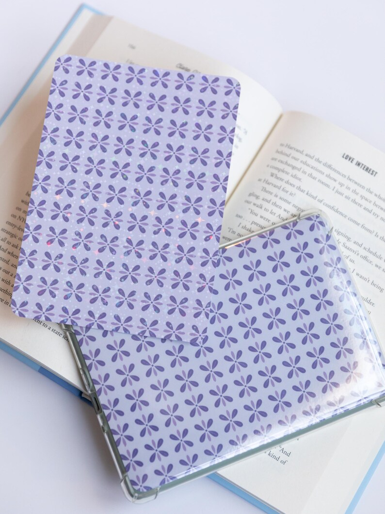 May include: A clear phone case and a matching card with a floral pattern in shades of purple rest on an open book. The case is transparent, showcasing the design beneath. The book's pages are visible, adding context to the scene.