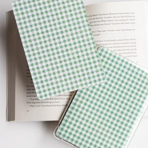 May include: Green and white gingham patterned phone case and bookmark. The phone case is clear with a green and white checkered design. The bookmark has the same pattern. A book is in the background.