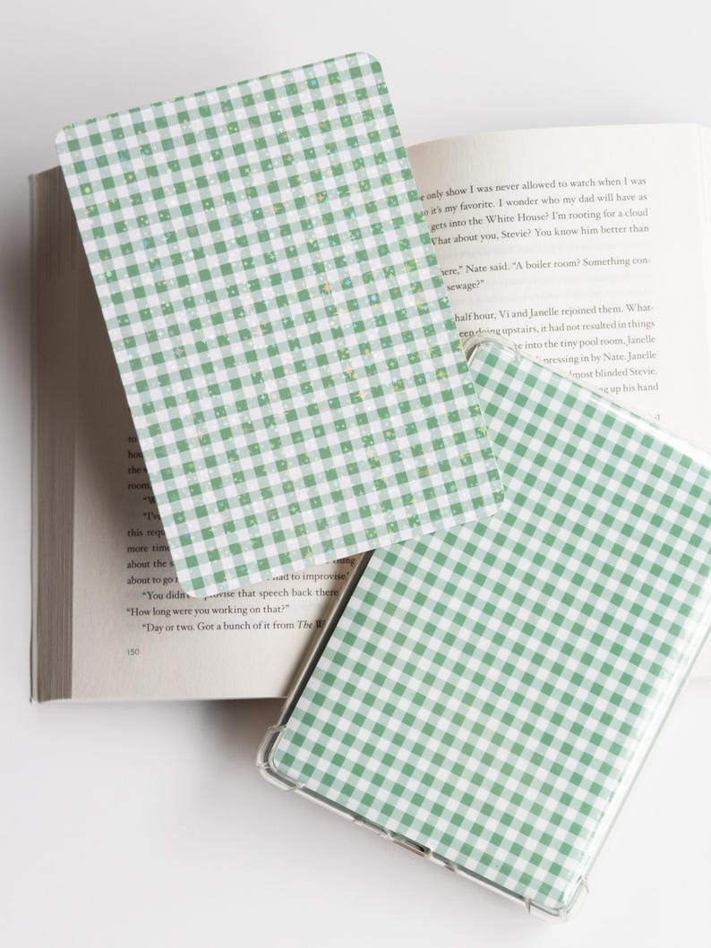 May include: Green and white gingham patterned phone case and bookmark. The phone case is clear with a green and white checkered design. The bookmark has the same pattern. A book is in the background.