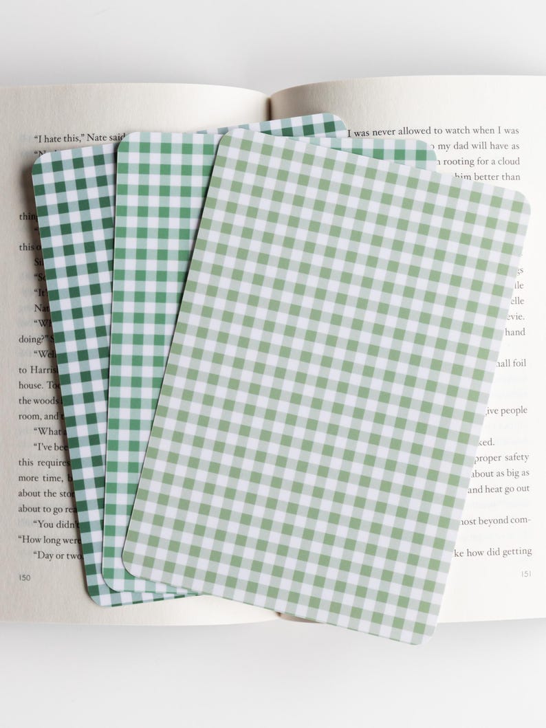 May include: Three rectangular cards with a green and white gingham pattern are fanned out on top of an open book. The cards are a light green color and have rounded corners. The book pages are visible in the background.