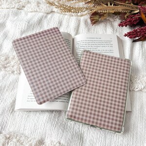 May include: Two rectangular e-reader or tablet cases featuring a dusty rose and white gingham check pattern. One case is placed flat on an open book, while the other is positioned slightly overlapping it, showing a clear protective edge.