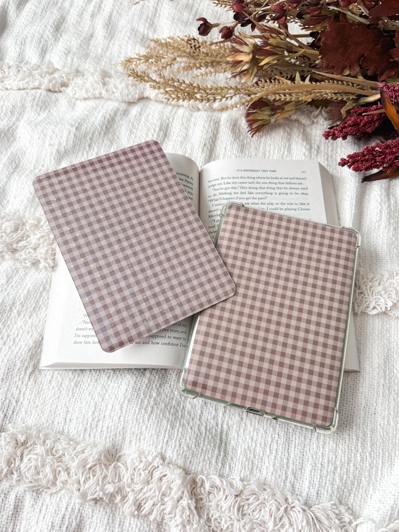 May include: Two rectangular e-reader or tablet cases featuring a dusty rose and white gingham check pattern. One case is placed flat on an open book, while the other is positioned slightly overlapping it, showing a clear protective edge.