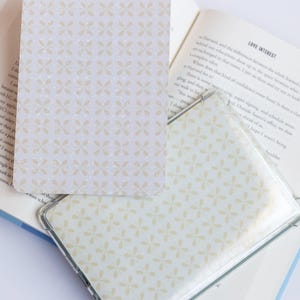 May include: A clear plastic case and a card with a light beige floral pattern. The card is rectangular with rounded corners. The pattern is repeated across the surface. The case is open, revealing the pattern.