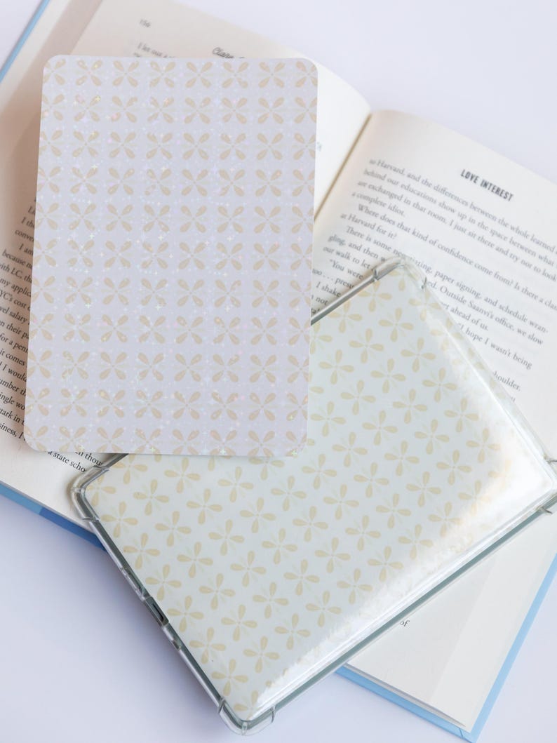May include: A clear plastic case and a card with a light beige floral pattern. The card is rectangular with rounded corners. The pattern is repeated across the surface. The case is open, revealing the pattern.
