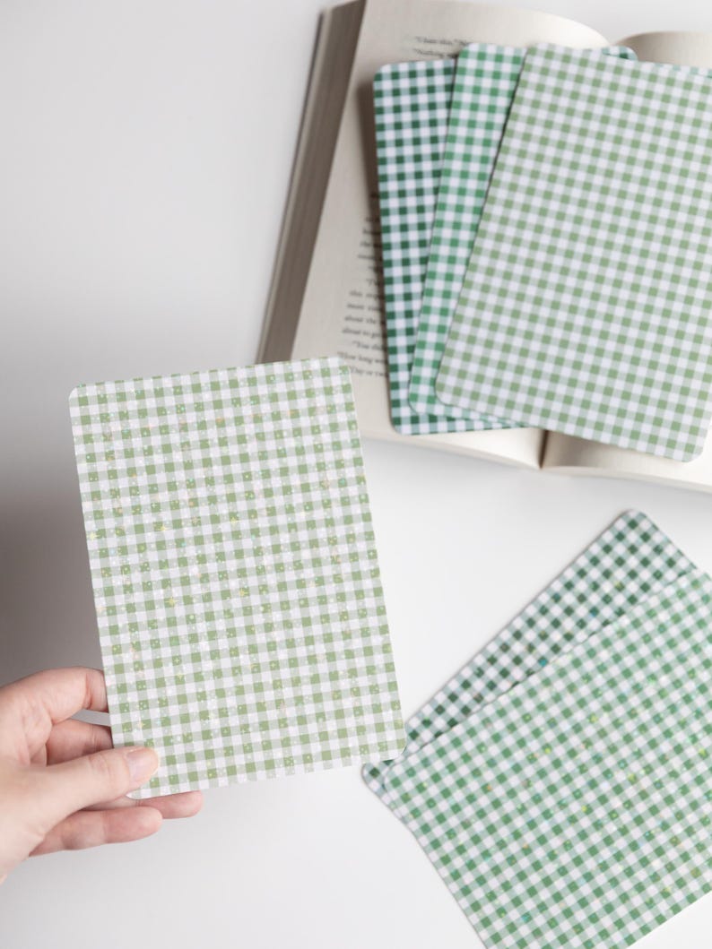 May include: A stack of green and white gingham patterned cards next to an open book. One card is being held by a hand. The cards have rounded corners and a light green and white checkered design. The book pages are visible in the background.