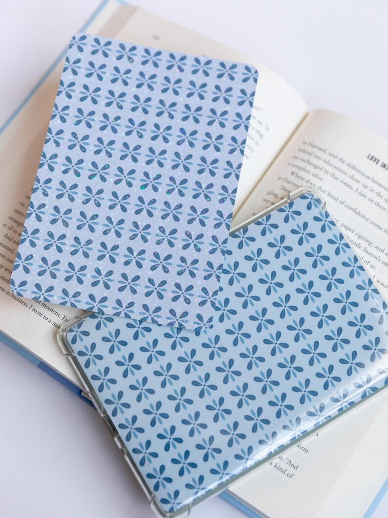 May include: Two blue patterned book covers with a floral design are displayed open on a book. The covers have a clear plastic sleeve. The book pages are visible, with text and a bookmark.