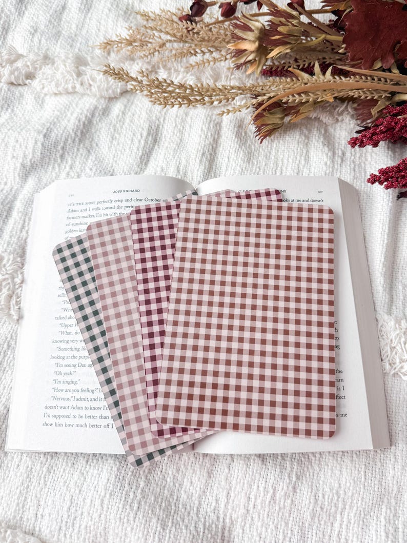 May include: A set of four rectangular bookmarks featuring a gingham check pattern in varying shades of brown, pink, and dark green. The bookmarks are arranged in a fan shape and are resting on the open pages of a printed book with visible text.