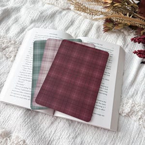 May include: Three rectangular bookmarks with rounded corners featuring plaid patterns in shades of deep burgundy, muted pink, and sage green. The bookmarks are layered on top of an open book with visible printed text on the white paper pages.
