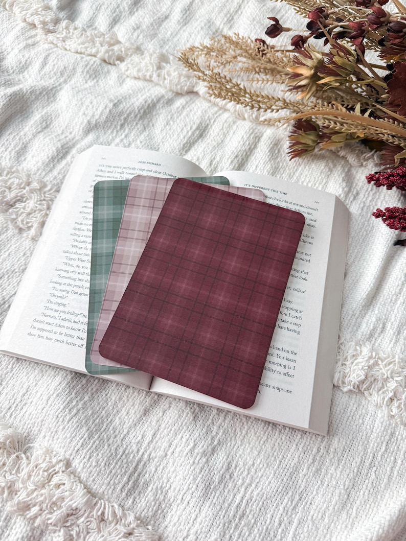 May include: Three rectangular bookmarks with rounded corners featuring plaid patterns in shades of deep burgundy, muted pink, and sage green. The bookmarks are layered on top of an open book with visible printed text on the white paper pages.