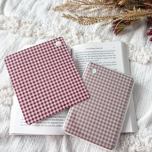 May include: Two square coasters featuring a red and white gingham check pattern. Each coaster has a small white circular accent in the corner. The coasters are positioned on top of an open book with visible printed text on the pages.